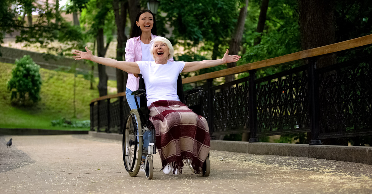 Long-Term Happiness. Picture of an elderly caucasian female in a wheel chair, being pushed by a young asian female.